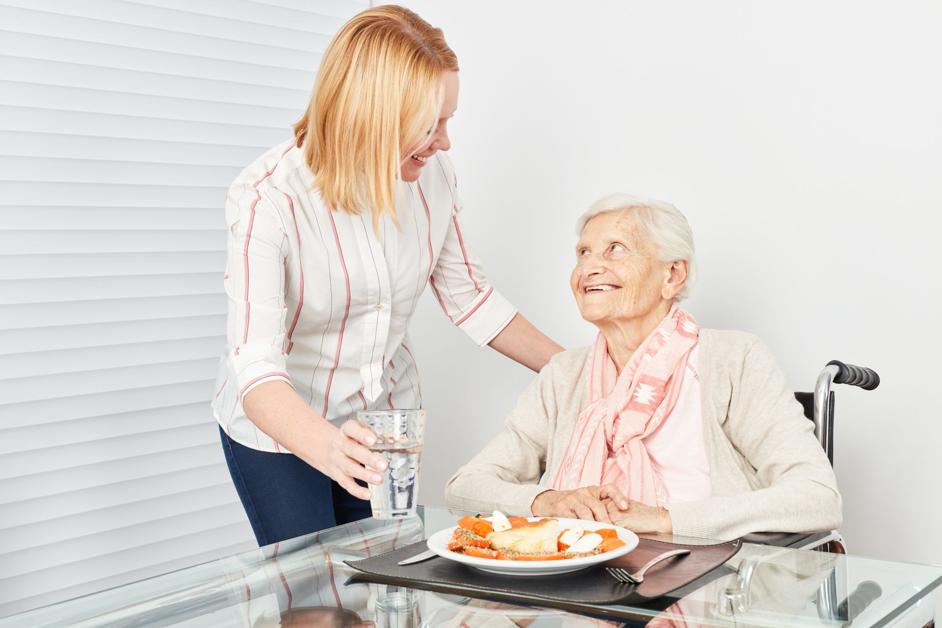 Pflegekraft reicht Seniorin ein Glas Wasser am Esstisch